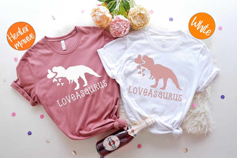 May include: Two t-shirts with a dinosaur graphic and the text "Loveasaurus". One shirt is white with a pink dinosaur and the other is a heather mauve color with a white dinosaur.