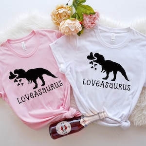 May include: Two pink and white t-shirts with a black silhouette of a dinosaur holding hearts in its mouth. The text "Loveasaurus" is printed on each shirt.