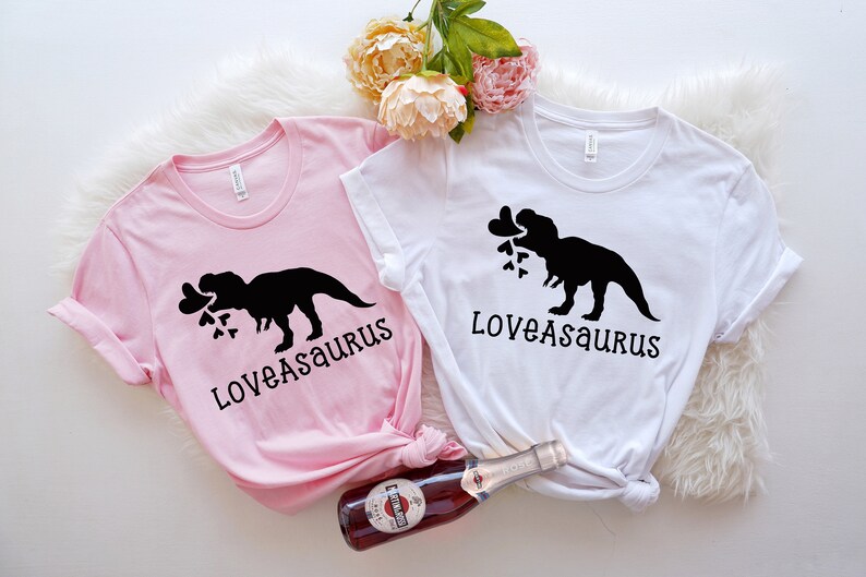 May include: Two pink and white t-shirts with a black silhouette of a dinosaur holding hearts in its mouth. The text "Loveasaurus" is printed on each shirt.