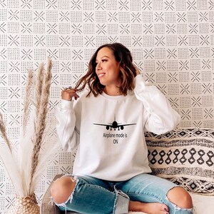 May include: A white sweatshirt with a black airplane graphic and the text "Airplane mode is ON". The model is wearing the sweatshirt and blue jeans.