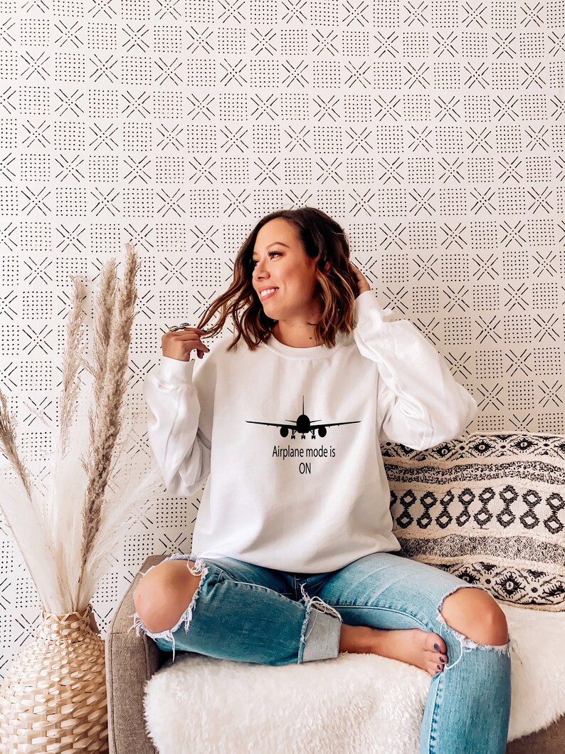 May include: A white sweatshirt with a black airplane graphic and the text "Airplane mode is ON". The model is wearing the sweatshirt and blue jeans.