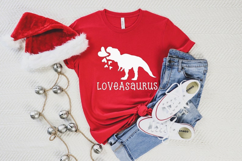 May include: Red t-shirt with a white dinosaur holding mistletoe and the text "Loveasaurus". The shirt is paired with blue jeans and white sneakers.