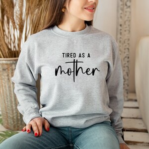 May include: A gray sweatshirt with the text "TIRED AS A MOTHER" printed on the front. The text is in black and has a cross in the middle of the word "mother".
