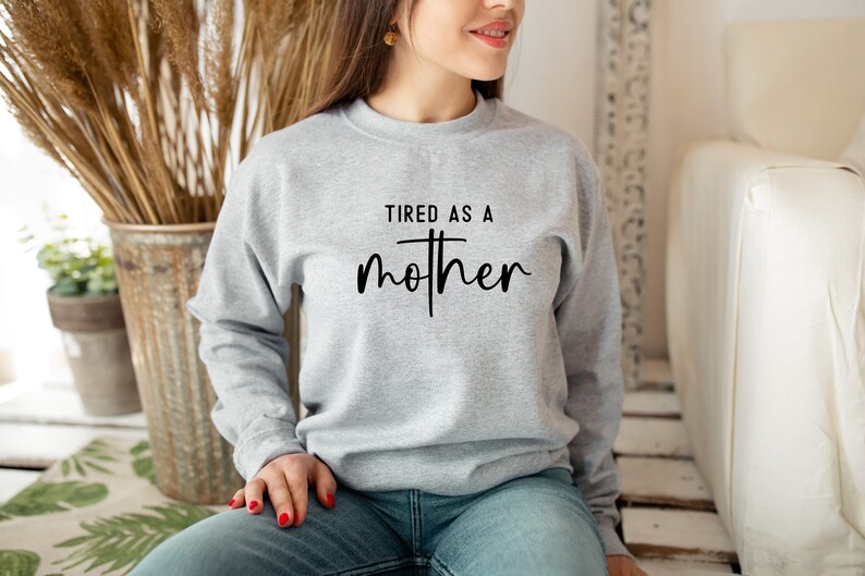 May include: A gray sweatshirt with the text "TIRED AS A MOTHER" printed on the front. The text is in black and has a cross in the middle of the word "mother".