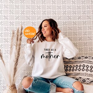 May include: White sweatshirt with the text "TIRED AS A MOTHER" in black lettering. The sweatshirt is being worn by a person sitting on a couch.