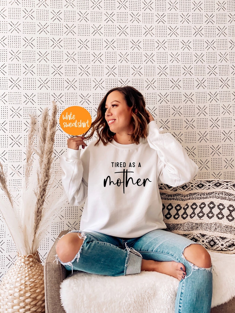 May include: White sweatshirt with the text "TIRED AS A MOTHER" in black lettering. The sweatshirt is being worn by a person sitting on a couch.