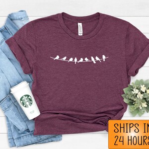 May include: A maroon t-shirt with a white graphic of a line of birds on a wire. The t-shirt is on a white wooden surface. The text "SHIPS IN 24 HOURS" is in orange on a white background.