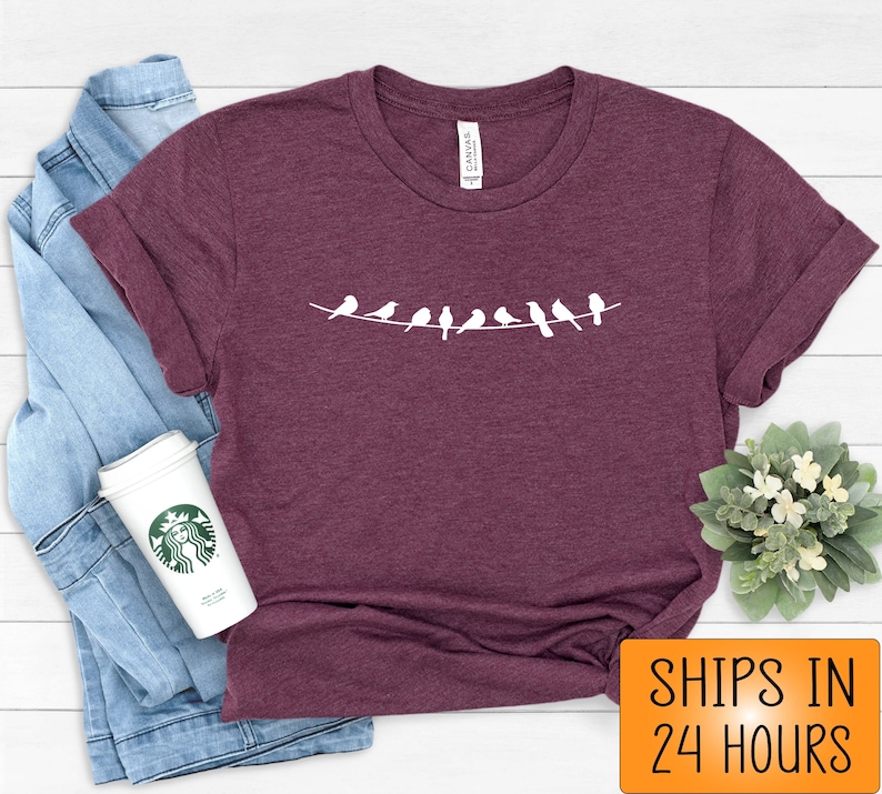 May include: A maroon t-shirt with a white graphic of a line of birds on a wire. The t-shirt is on a white wooden surface. The text "SHIPS IN 24 HOURS" is in orange on a white background.