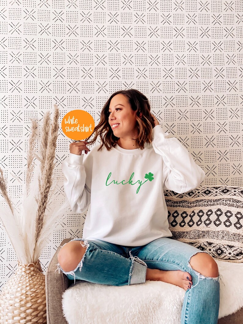 May include: A white sweatshirt with the word "lucky" written in green cursive with a green four-leaf clover next to it. The sweatshirt is being worn by a person sitting on a couch.