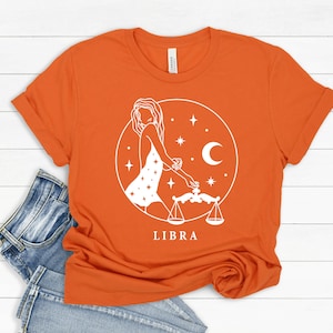May include: Orange t-shirt with a white graphic of a woman with a Libra zodiac symbol and the word "LIBRA" printed below.