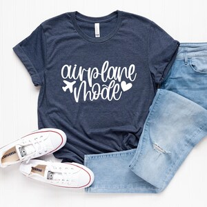 May include: A navy blue t-shirt with white text that reads "airplane mode" with a small airplane and heart graphic. The shirt is paired with blue jeans and white sneakers.