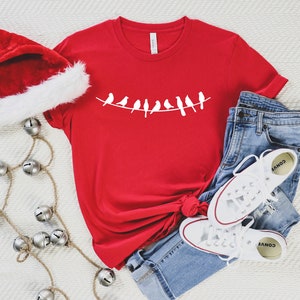 May include: Red t-shirt with a white design of birds on a wire. The shirt is paired with blue jeans and white sneakers.
