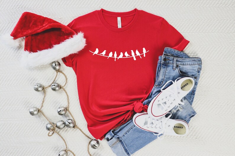 May include: Red t-shirt with a white design of birds on a wire. The shirt is paired with blue jeans and white sneakers.