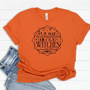 May include: Orange t-shirt with the text "We are the granddaughters of the witches you couldn't burn" in a black geometric design. The shirt is folded, with a pair of blue jeans visible in the background.