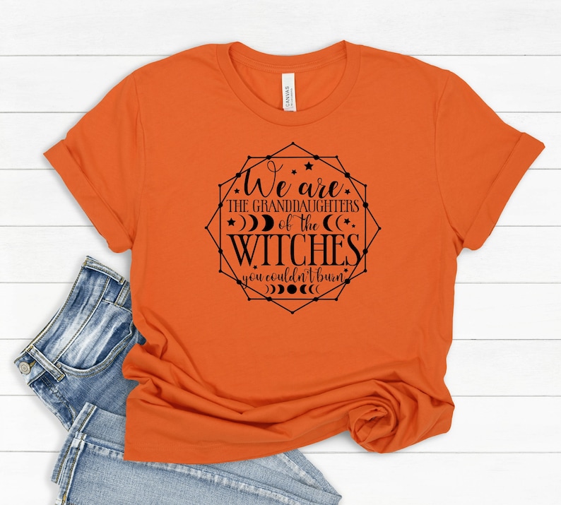 May include: Orange t-shirt with the text "We are the granddaughters of the witches you couldn't burn" in a black geometric design. The shirt is folded, with a pair of blue jeans visible in the background.