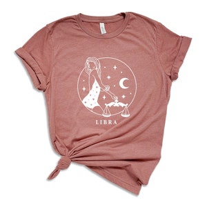 May include: A dusty rose t-shirt with a white graphic of a Libra zodiac sign. The graphic features a woman holding a scale and a crescent moon.