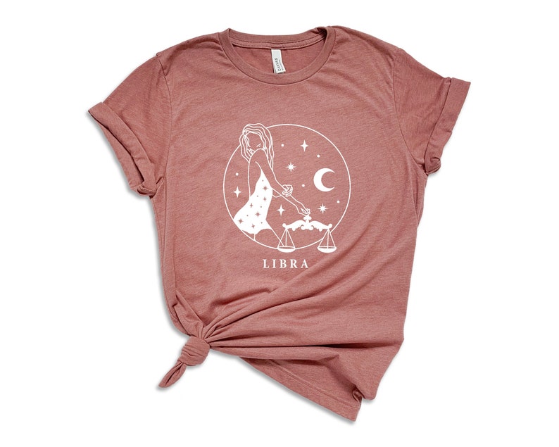 May include: A dusty rose t-shirt with a white graphic of a Libra zodiac sign. The graphic features a woman holding a scale and a crescent moon.