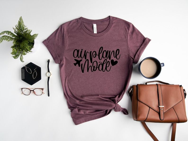 May include: A mauve colored t-shirt with the text "airplane mode" in black lettering. The shirt is folded and tied in a knot. A small airplane icon and two hearts are also included in the design.