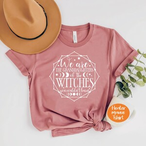 May include: A heather mauve t-shirt with rolled sleeves and a graphic that reads "We are the granddaughters of the witches you couldn't burn." A tan hat and a sprig of greenery are next to the shirt.