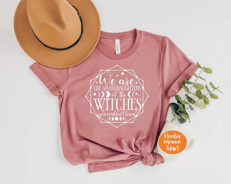 May include: A heather mauve t-shirt with rolled sleeves and a graphic that reads "We are the granddaughters of the witches you couldn't burn." A tan hat and a sprig of greenery are next to the shirt.