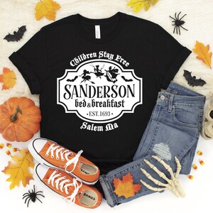 May include: Black t-shirt with a white graphic of a sign that says "Sanderson Bed & Breakfast - Est. 1693 - Salem Ma" with a witch and bats flying around it. The text above the sign says "Children Stay Free".