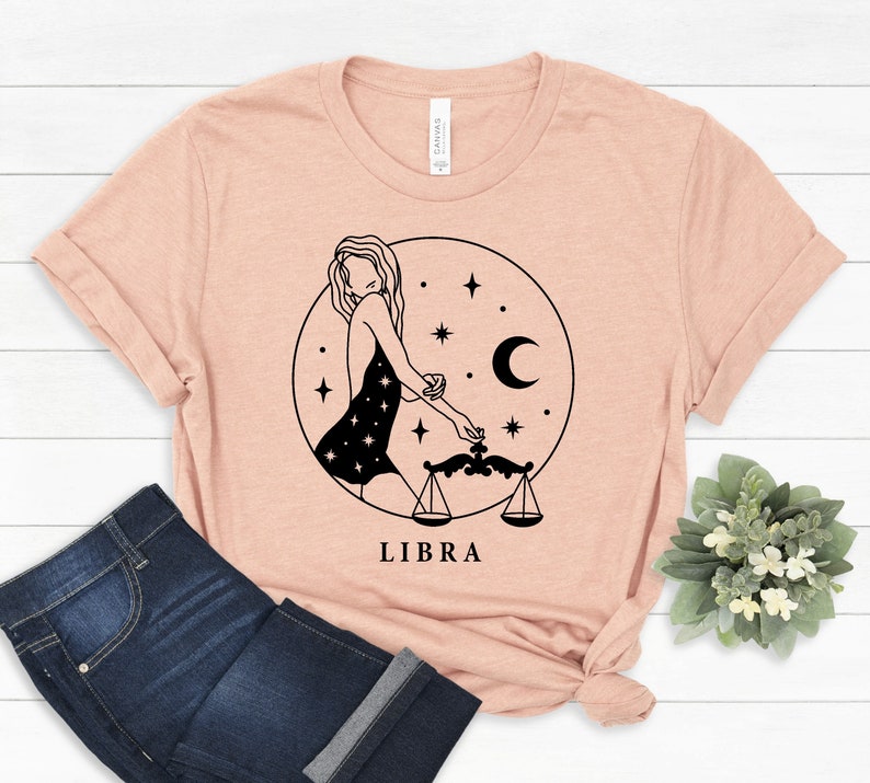 May include: A peach-colored t-shirt with a black line drawing of a woman holding a scale, a crescent moon, and stars. The text "LIBRA" is printed below the image.