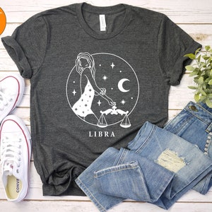 May include: A dark heather grey t-shirt with a white graphic of a woman holding a scale, a crescent moon, and stars. The text "LIBRA" is printed below the graphic.