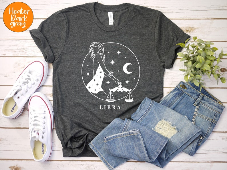 May include: A dark heather grey t-shirt with a white graphic of a woman holding a scale, a crescent moon, and stars. The text "LIBRA" is printed below the graphic.
