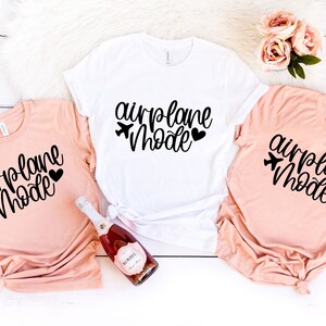 May include: Three t-shirts with the text "airplane mode" in black script with a small airplane icon and heart. Two shirts are peach colored and one is white.