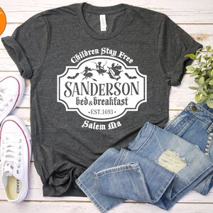 May include: A dark heather gray t-shirt with a white graphic of a vintage sign that reads "Children Stay Free Sanderson Bed & Breakfast Est. 1693 Salem Ma". The sign features a silhouette of three witches flying on broomsticks.
