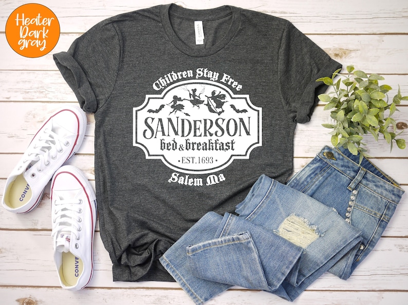 May include: A dark heather gray t-shirt with a white graphic of a vintage sign that reads "Children Stay Free Sanderson Bed & Breakfast Est. 1693 Salem Ma". The sign features a silhouette of three witches flying on broomsticks.