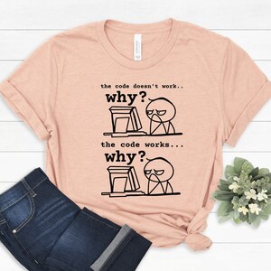 May include: A peach-colored t-shirt with a black and white illustration of a stick figure at a computer. The text on the shirt reads "the code doesn't work... why?" and "the code works... why?"