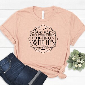 May include: A light peach-colored t-shirt with the text "We are the granddaughters of the witches you couldn't burn" in a black geometric design. The shirt is paired with dark blue jeans.