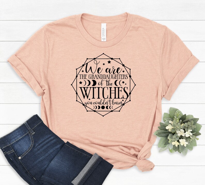 May include: A light peach-colored t-shirt with the text "We are the granddaughters of the witches you couldn't burn" in a black geometric design. The shirt is paired with dark blue jeans.