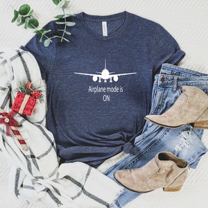 May include: A navy blue t-shirt with a white airplane graphic and the text "Airplane mode is ON". The shirt is laid out on a white surface with a plaid blanket, a pair of jeans, and brown ankle boots.