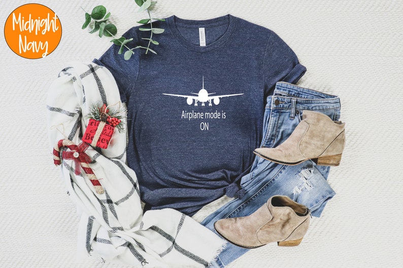 May include: A navy blue t-shirt with a white airplane graphic and the text "Airplane mode is ON". The shirt is laid out on a white surface with a plaid blanket, a pair of jeans, and brown ankle boots.