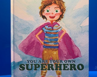 May include: A book cover with a watercolor illustration of a child wearing a pink cape and a striped shirt. The text on the cover reads "You are your own Superhero" by Adam Mills. Art by Branislav Sosic Lettered by Daniel Powers.