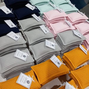 May include: Multiple stacks of rectangular heat and cold packs made of waffle-textured fabric in navy blue, mint green, light pink, gray, and mustard yellow. Each pack features a white paper tag tied with string labeled Multipurpose Heat/Cold Pack.
