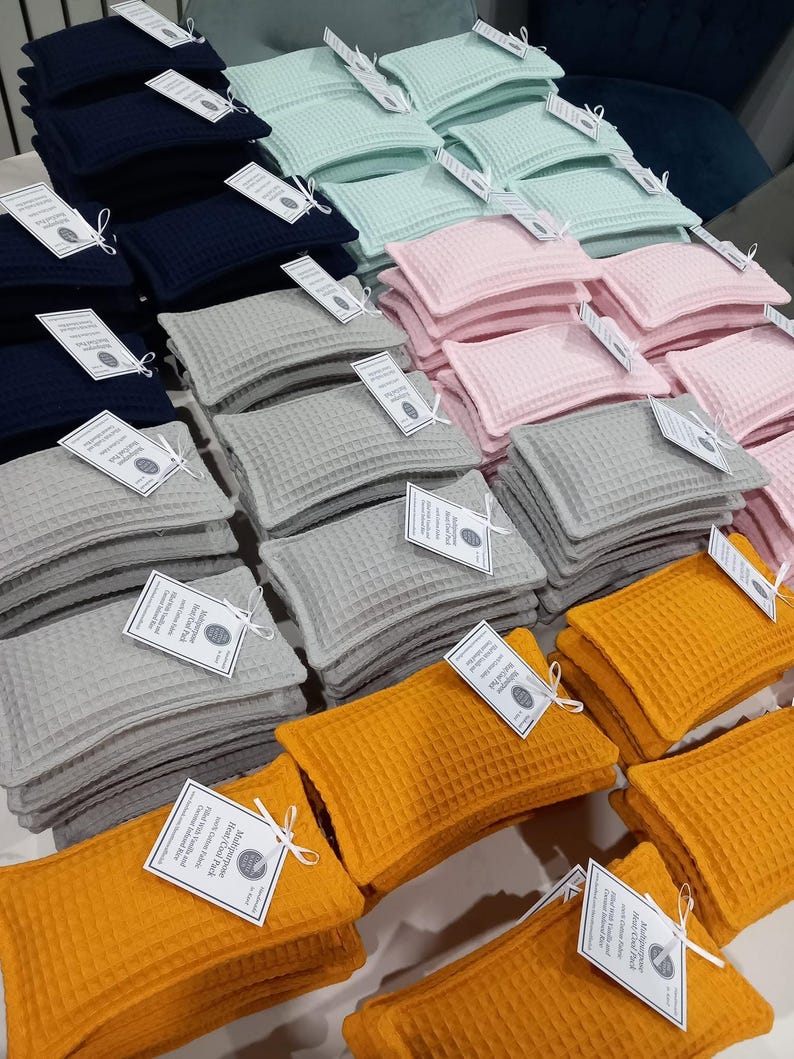 May include: Multiple stacks of rectangular heat and cold packs made of waffle-textured fabric in navy blue, mint green, light pink, gray, and mustard yellow. Each pack features a white paper tag tied with string labeled Multipurpose Heat/Cold Pack.