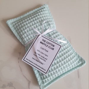 May include: A rectangular light mint green waffle-weave fabric cold pack tied with a thin white ribbon. A white tag attached to the ribbon reads: THE COTTON WAFFLE CLUB, Cold Pack, 100% Cotton Fabric, Filled With Vanilla and Coconut Infused Rice.