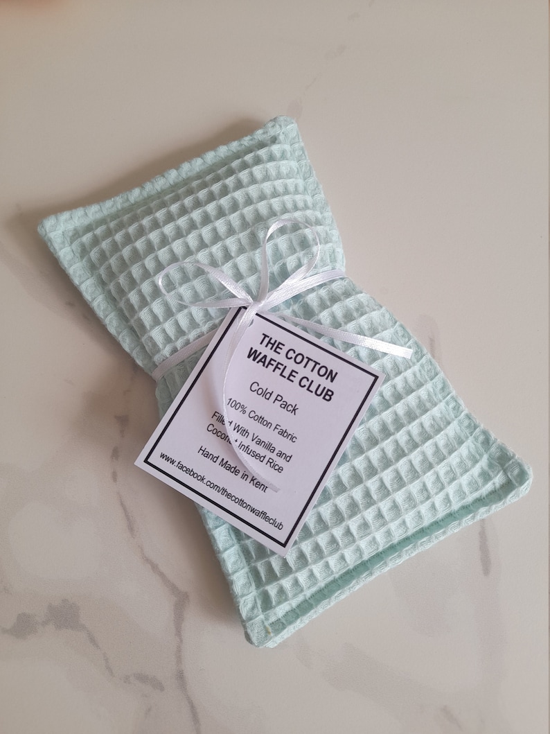 May include: A rectangular light mint green waffle-weave fabric cold pack tied with a thin white ribbon. A white tag attached to the ribbon reads: THE COTTON WAFFLE CLUB, Cold Pack, 100% Cotton Fabric, Filled With Vanilla and Coconut Infused Rice.