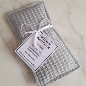 May include: A rectangular light gray cold pack made of waffle-textured cotton fabric. A white ribbon ties a square tag to the pack. The tag reads: THE COTTON WAFFLE CLUB, Cold Pack, 100% Cotton Fabric, Filled With Vanilla and Coconut Infused Rice, Hand Made in Kent, www.facebook.com/thecottonwaffleclub.