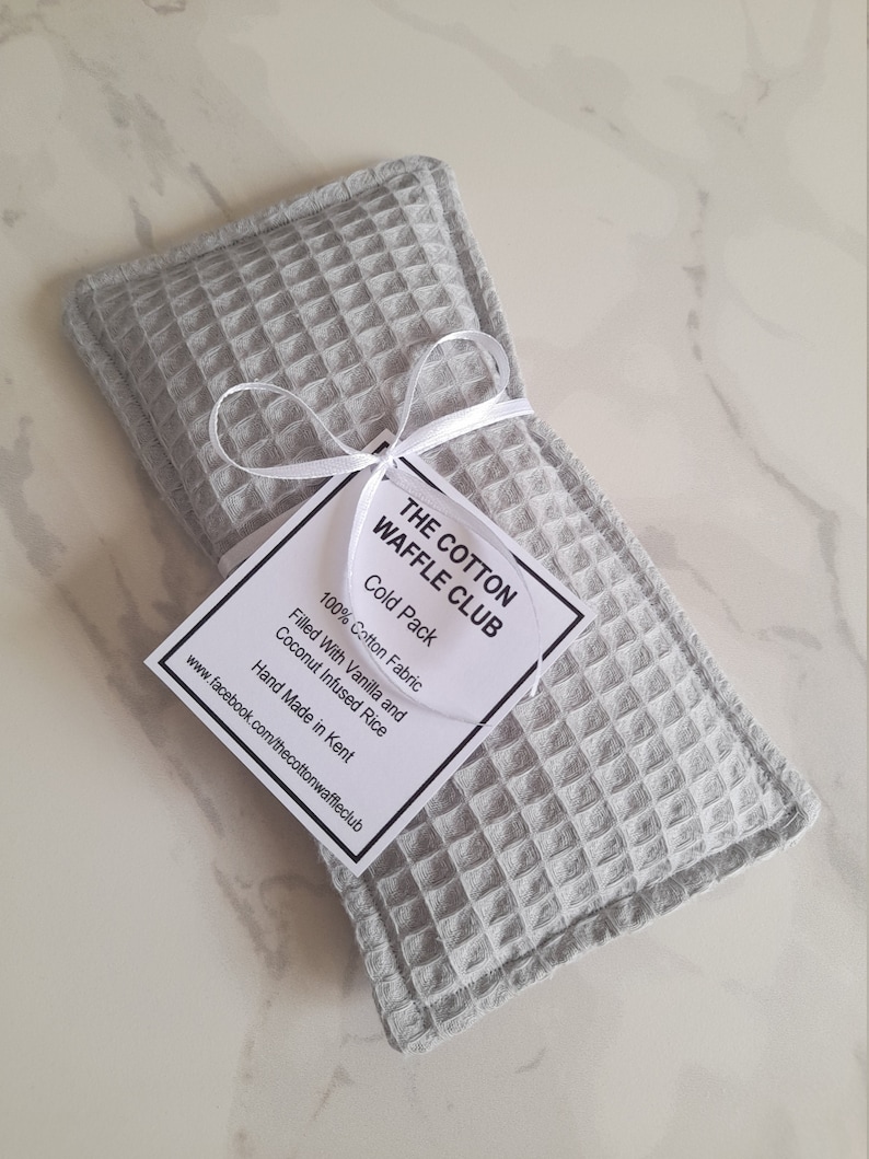 May include: A rectangular light gray cold pack made of waffle-textured cotton fabric. A white ribbon ties a square tag to the pack. The tag reads: THE COTTON WAFFLE CLUB, Cold Pack, 100% Cotton Fabric, Filled With Vanilla and Coconut Infused Rice, Hand Made in Kent, www.facebook.com/thecottonwaffleclub.