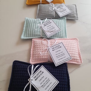 May include: Five rectangular cold packs made of waffle-textured cotton fabric in mustard yellow, light gray, mint green, pale pink, and navy blue. Each pack is tied with a white ribbon and a tag labeled The Cotton Waffle Club, Cold Pack, 100% Cotton Fabric.