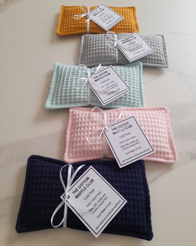 May include: Five rectangular cold packs made of waffle-textured cotton fabric in mustard yellow, light gray, mint green, pale pink, and navy blue. Each pack is tied with a white ribbon and a tag labeled The Cotton Waffle Club, Cold Pack, 100% Cotton Fabric.