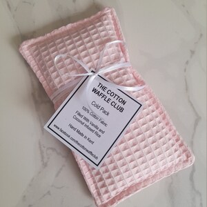 May include: A rectangular light pink cold pack made of waffle-textured cotton fabric. A white ribbon ties a square tag to the center, which reads: THE COTTON WAFFLE CLUB, Cold Pack, 100% Cotton Fabric, Filled With Vanilla and Coconut Infused Rice.