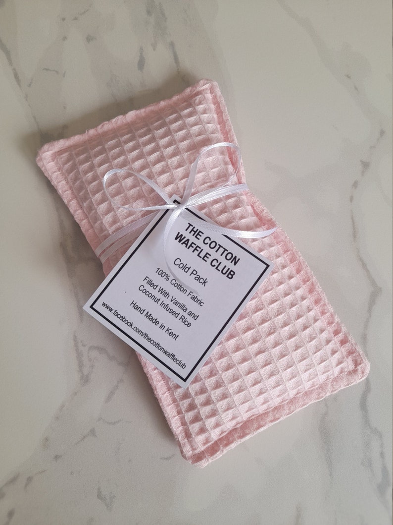 May include: A rectangular light pink cold pack made of waffle-textured cotton fabric. A white ribbon ties a square tag to the center, which reads: THE COTTON WAFFLE CLUB, Cold Pack, 100% Cotton Fabric, Filled With Vanilla and Coconut Infused Rice.