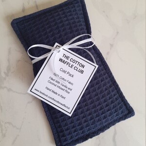 May include: A rectangular navy blue cold pack made of textured waffle-weave cotton fabric. A white tag tied with a white ribbon reads: THE COTTON WAFFLE CLUB, Cold Pack, 100% Cotton Fabric, Filled With Vanilla and Coconut Infused Rice, Hand Made in Kent.