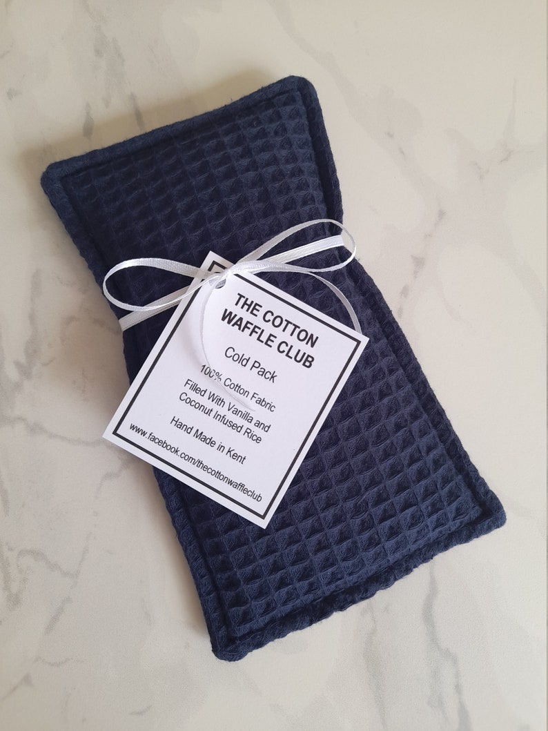 May include: A rectangular navy blue cold pack made of textured waffle-weave cotton fabric. A white tag tied with a white ribbon reads: THE COTTON WAFFLE CLUB, Cold Pack, 100% Cotton Fabric, Filled With Vanilla and Coconut Infused Rice, Hand Made in Kent.