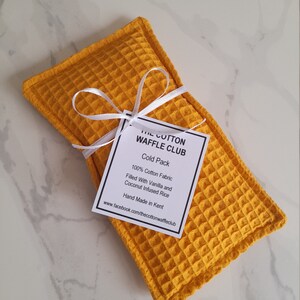 May include: A rectangular mustard yellow cold pack made of waffle-textured cotton fabric. A white ribbon is tied around the center, holding a white tag that reads: THE COTTON WAFFLE CLUB, Cold Pack, 100% Cotton Fabric, Filled With Vanilla and Coconut Infused Rice, Hand Made in Kent, www.facebook.com/thecottonwaffleclub.